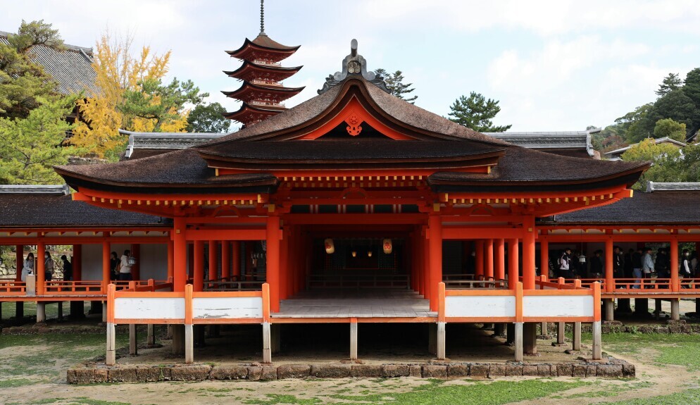 A traditional Japanese shrine with vibrant orange pillars and a curved roof stands on a stone platform. A pagoda rises in the background, surrounded by green and yellow trees under a partly cloudy sky.