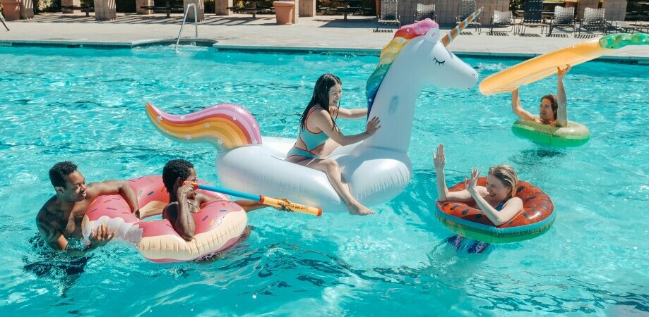 Four people are enjoying a swimming pool. One person is on a unicorn float, another on a watermelon float, while two more are on a donut float. One person holds an inflatable toy. The scene is bright and cheerful.