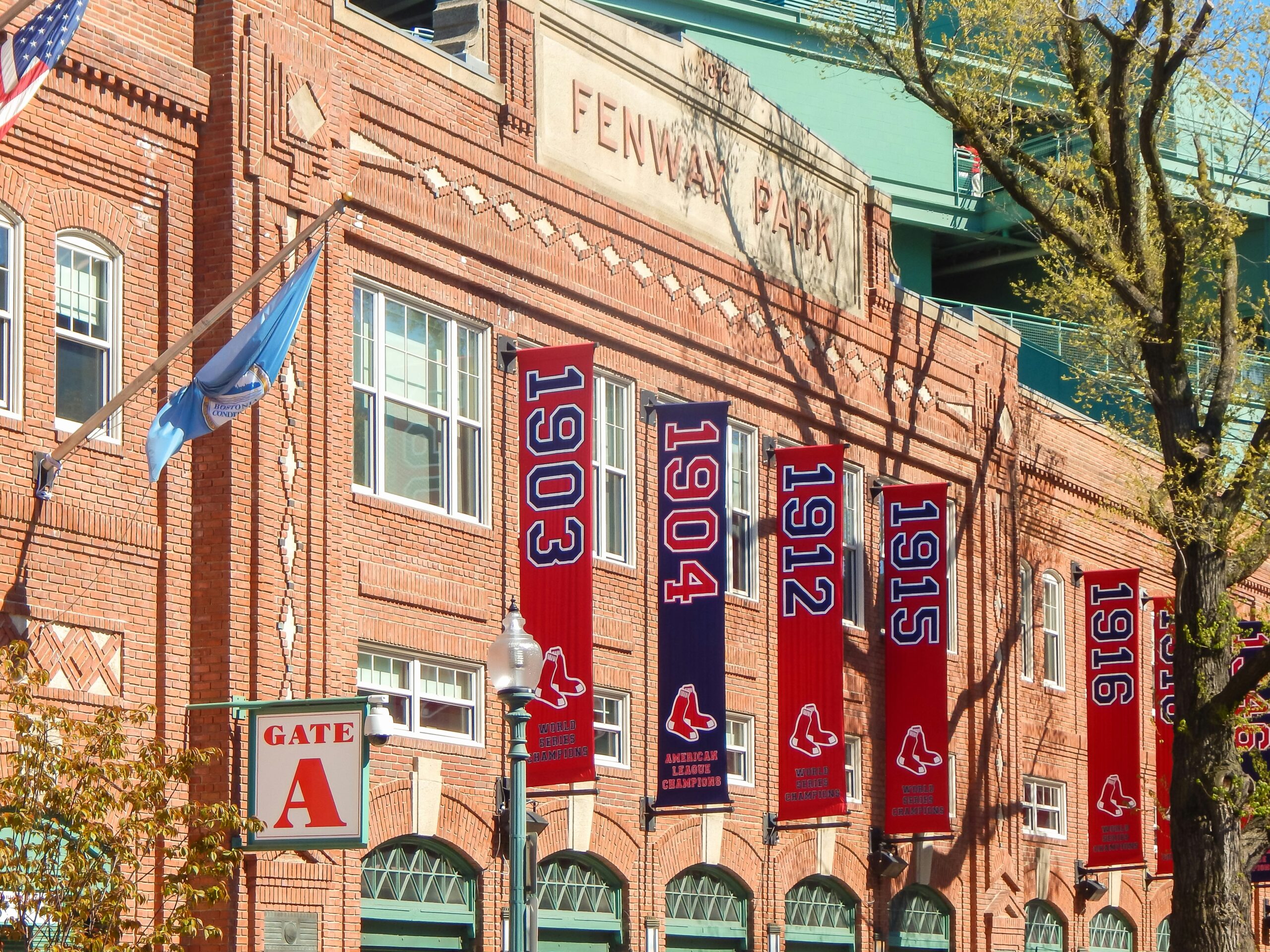 Brick exterior of Fenway Park, featuring multiple red and blue banners displaying years 1903, 1904, 1912, 1915, and 1916 with a baseball logo. The Gate A sign and an American flag are visible. Trees frame the scene.