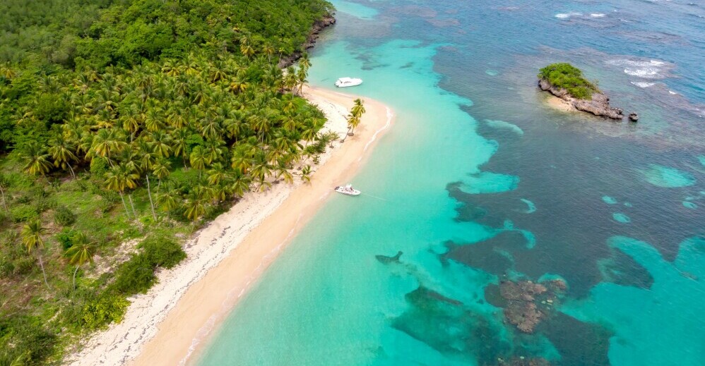 Aerial view of a tropical beach with lush green palm trees, golden sand, and turquoise water. Two boats are anchored near the shore. Small rocky land formations are visible in the water.