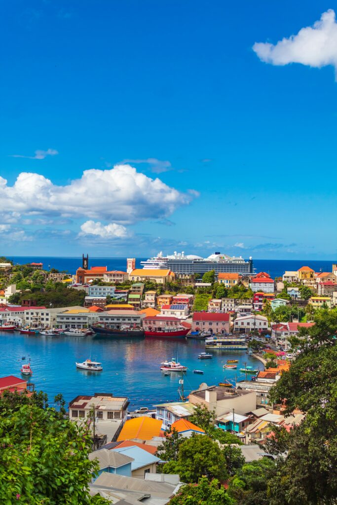 A vibrant coastal town in Grenada with colorful buildings next to a calm, blue bay. Boats are docked in the water, and a large cruise ship is in the background. The sky is bright blue with scattered white clouds.