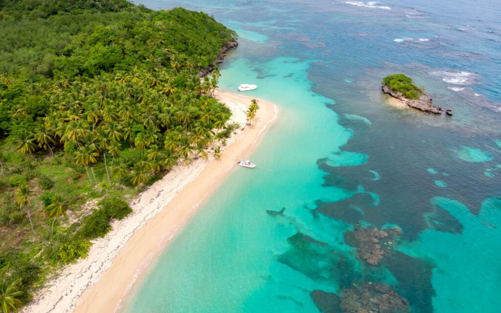 Aerial view of a tropical beach in Dominican Republic with golden sand and turquoise waters. Lush green palm trees line the shore. Two small boats are anchored near the waters edge. A rocky outcrop is visible in the ocean.