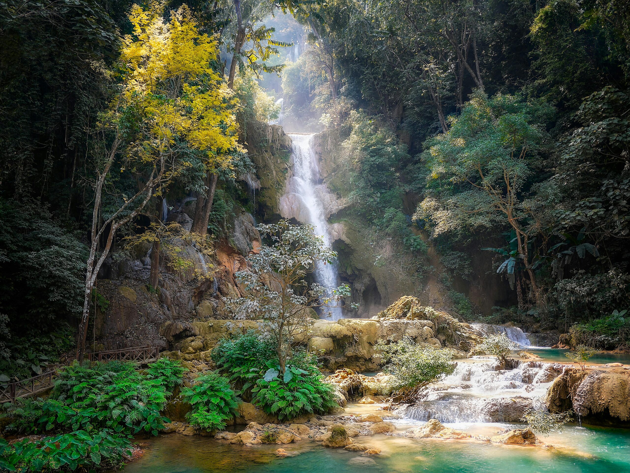 A cascading waterfall flows down terraced rocks surrounded by lush greenery and trees. Sunlight filters through the dense forest, illuminating the vibrant foliage and creating a serene atmosphere. A wooden railing lines a pathway on the left.