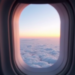 View from an airplane window showing a vibrant sunset above a sea of fluffy clouds. The sky transitions from deep blue to warm orange and pink hues near the horizon.