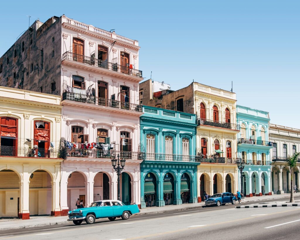Street view of colorful colonial buildings in Cuba with distinct arches and balconies. A vintage blue car is parked in front, and clothes hang on a line on one of the balconies. The sky is clear and blue, creating a vibrant and picturesque scene.