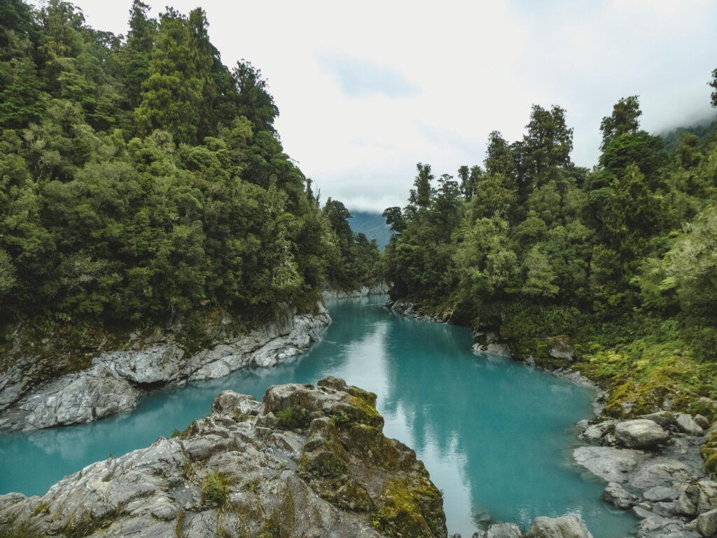 Scenic view of a turquoise river winding through lush green rainforest and rocky cliffs, under an overcast sky in a remote area of New Zealand