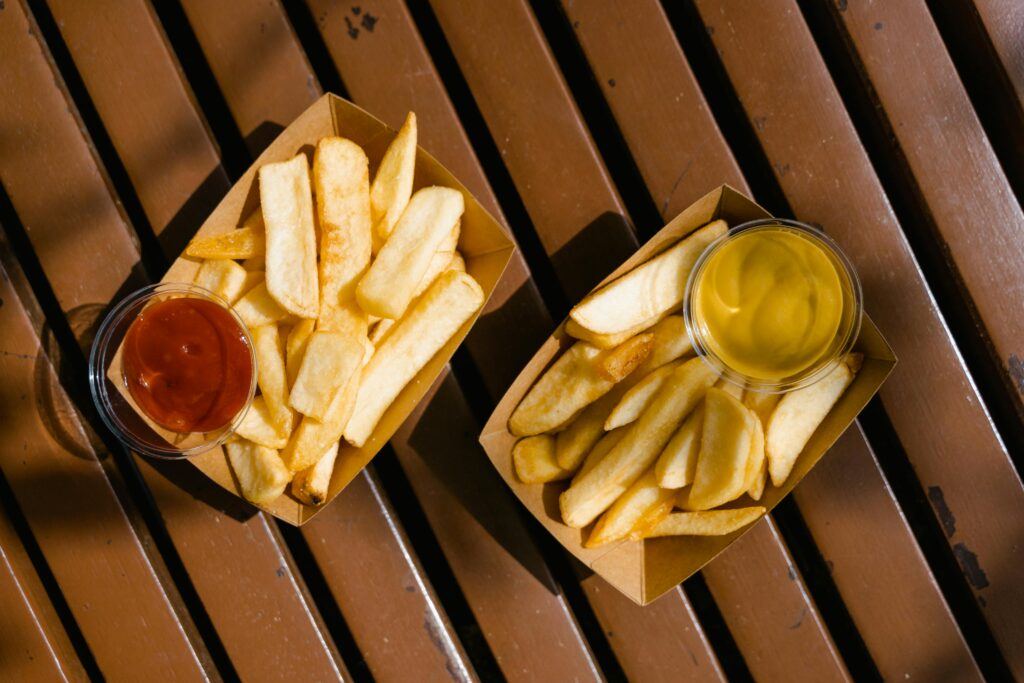 Two baskets of fries on top of a picnic table. One with mustard and one with ketchup.